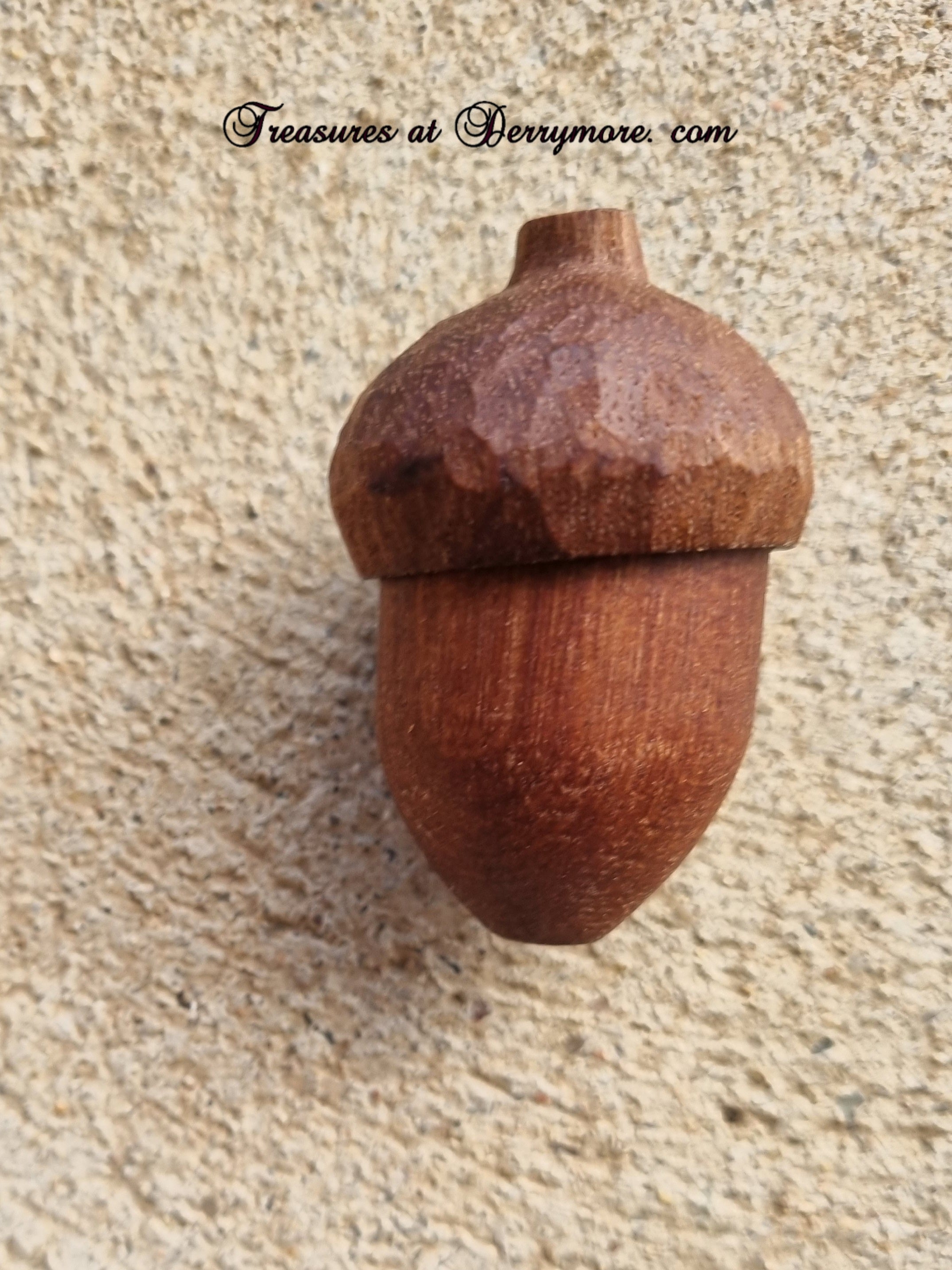 A carved wooden acorn ornament with a rich reddish-brown finish, mounted on a textured beige stucco wall. A watermark reading 'Treasures at Derrymore.