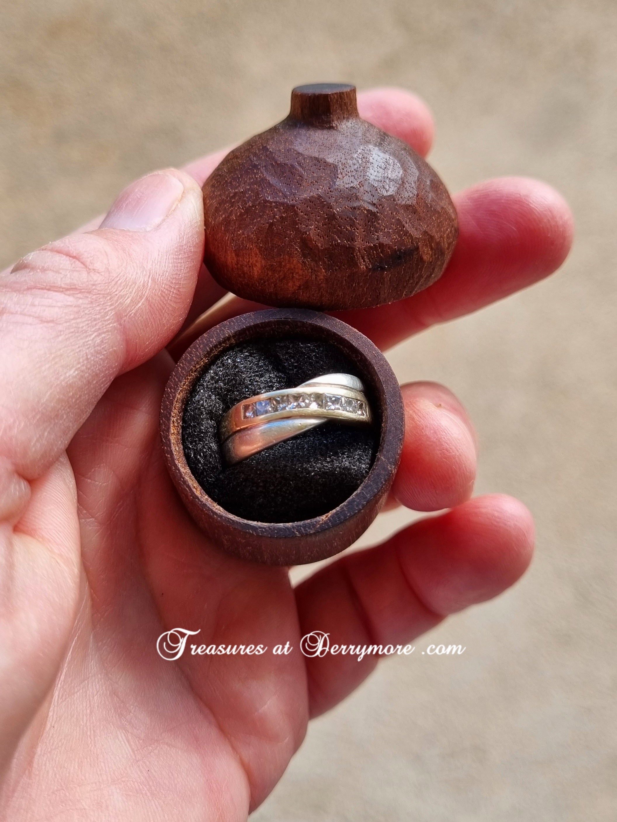 Hand carved acorn ring box. hand holds an open round wooden ring box with a dark cushion, displaying a silver band ring with a row of small clear stones. The wooden lid rests above the box against a soft beige background.