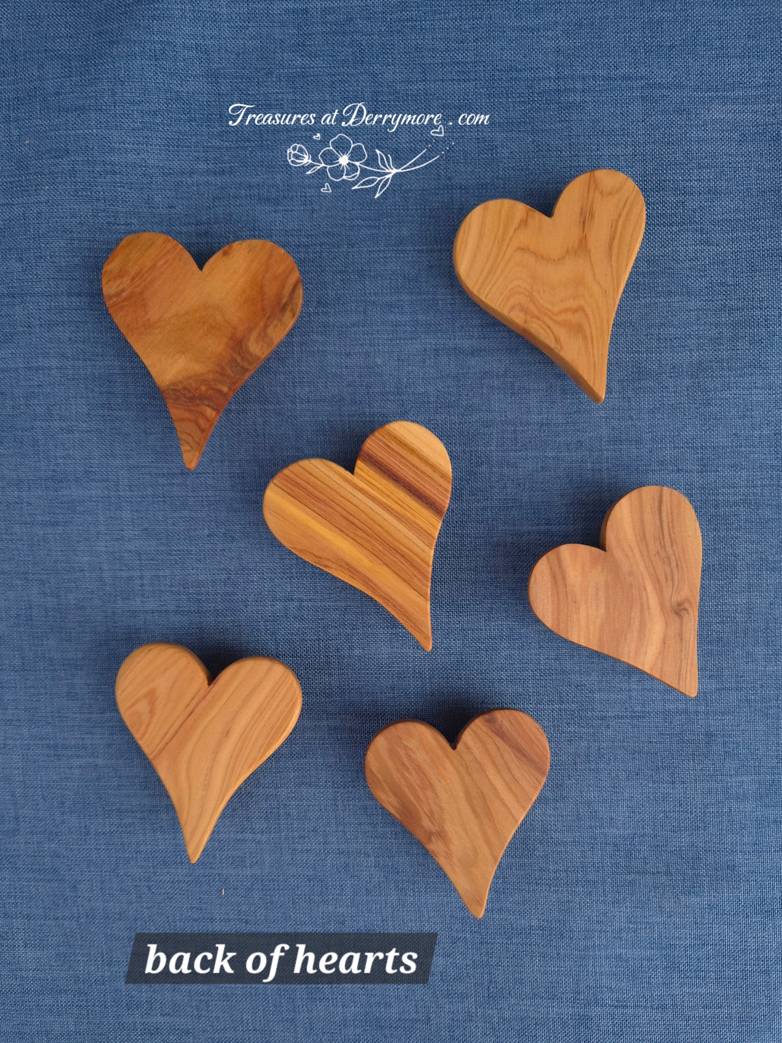 Six heart-shaped wooden pieces with visible wood grain patterns, arranged on a textured blue fabric background. White text at the top reads 'Treasures at Derrymere.