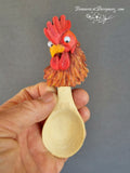 Wooden spoon with a rooster head decoration held by a hand against a gray background.