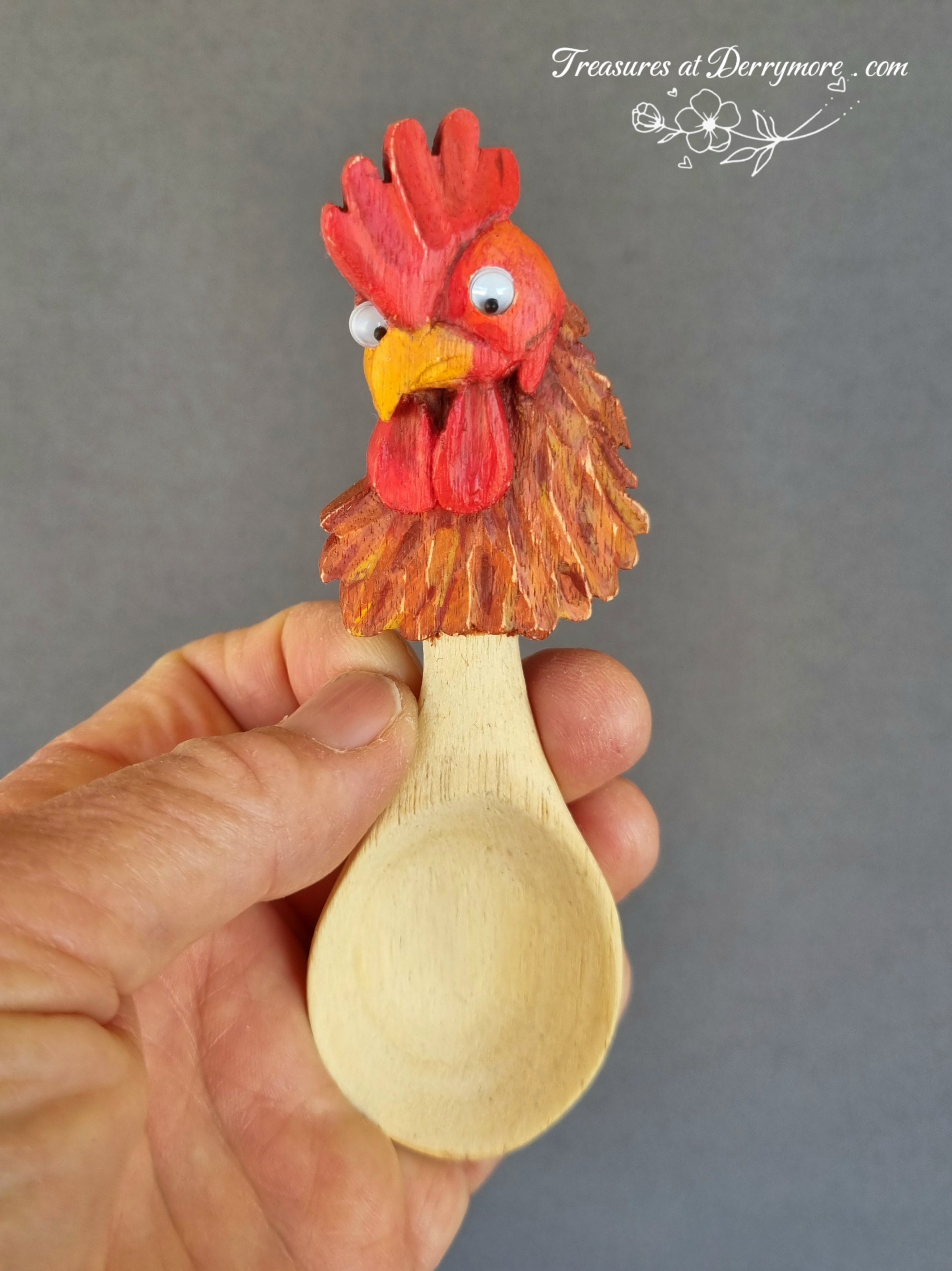 Wooden spoon with a rooster head decoration held by a hand against a gray background.