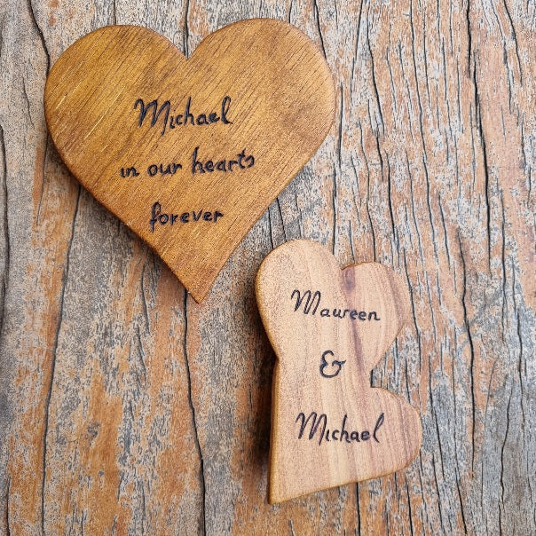 Two pyrography burnt wooden heart-shaped keepsakes on a weathered wood background. One larger heart is light brown wood with the engraved text 'Michael in our hearts forever', and one smaller heart is a medium tan wood with the engraved text