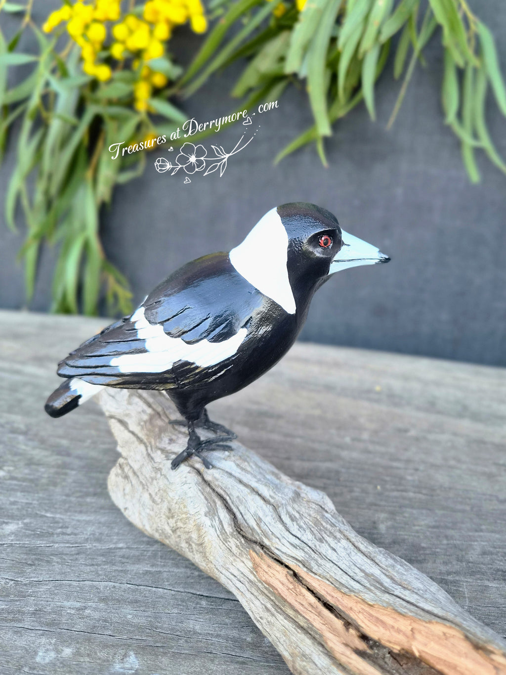 A hand-carved wooden Australian magpie with a black and white pattern, perched on a natural fallen piece of tree branch 