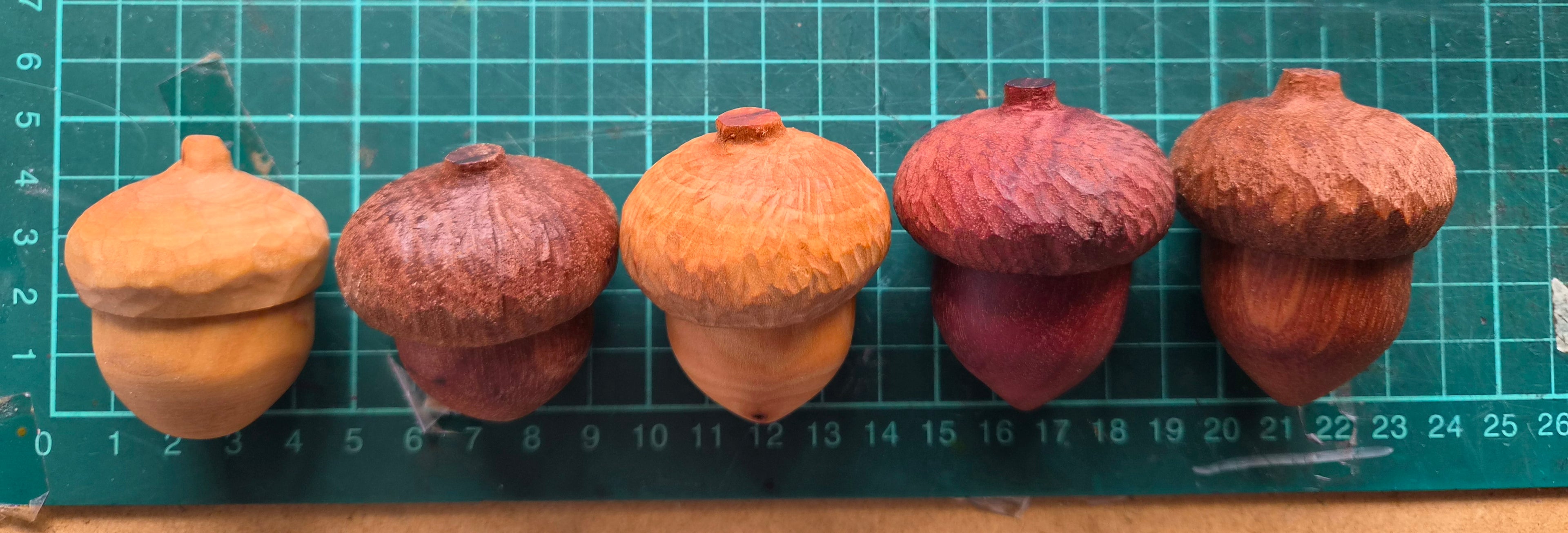 Five hand carved wooden acorn ring boxes in various shades of brown and reddish-brown, arranged in a row on a green cutting mat with grid lines and measurement markings.